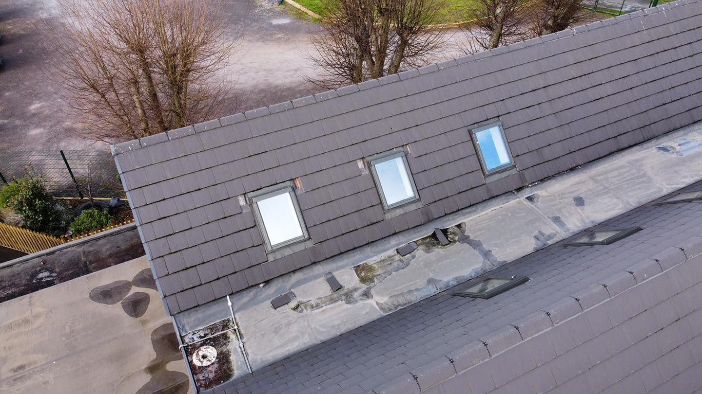 Aerial view of a grayish tiled roof with three skylights and a flat section. The flat part has some water stains and patches. Nearby trees lack leaves, indicating a winter or late fall season. Surrounding the area are parts of a parking lot and green space.