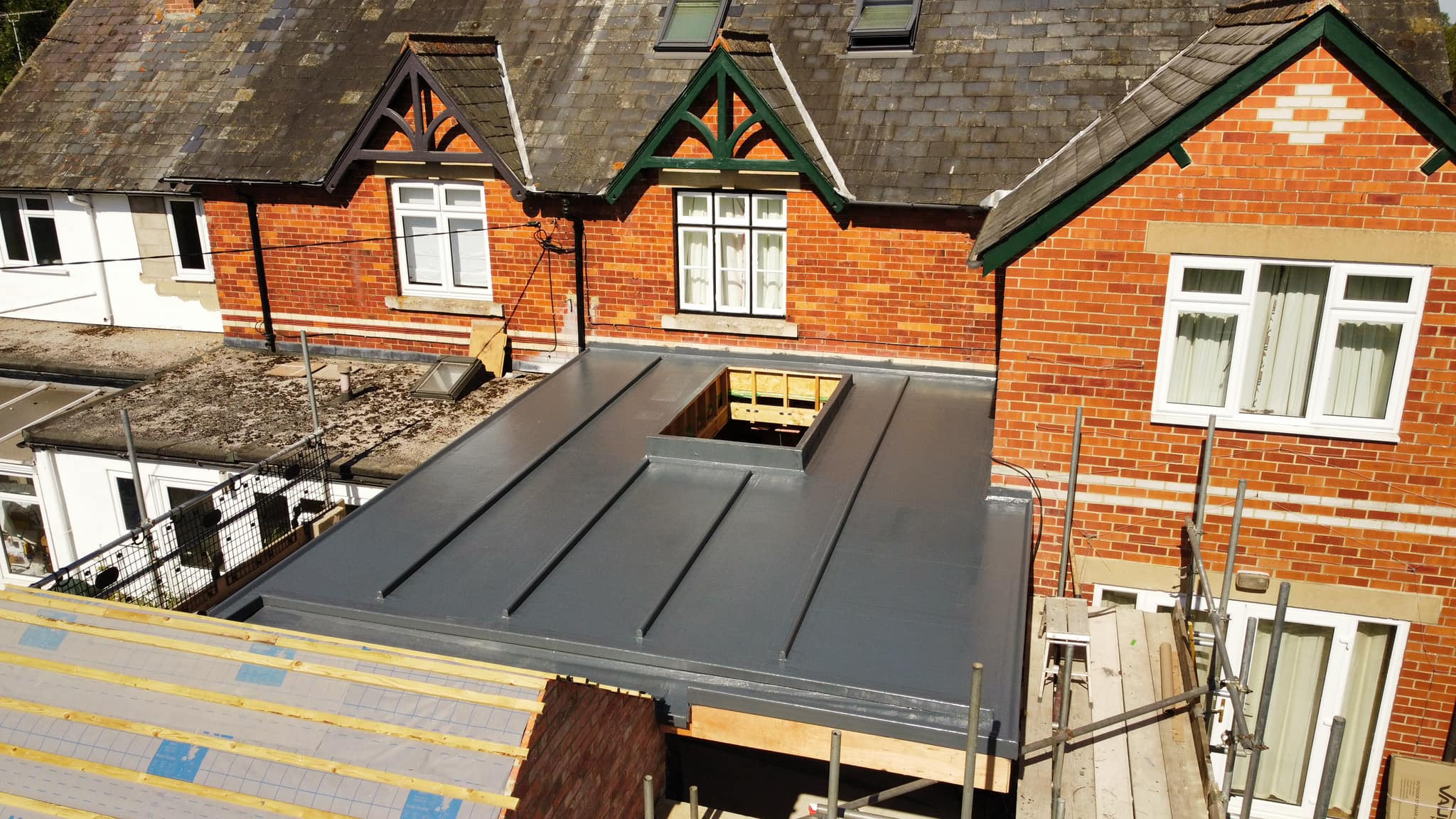 A rooftop view of a construction site with a partially completed flat roof extension connecting to a brick house. Scaffolding surrounds the site, and there's an unfinished section in the center of the new roof, likely for a skylight or ventilation.