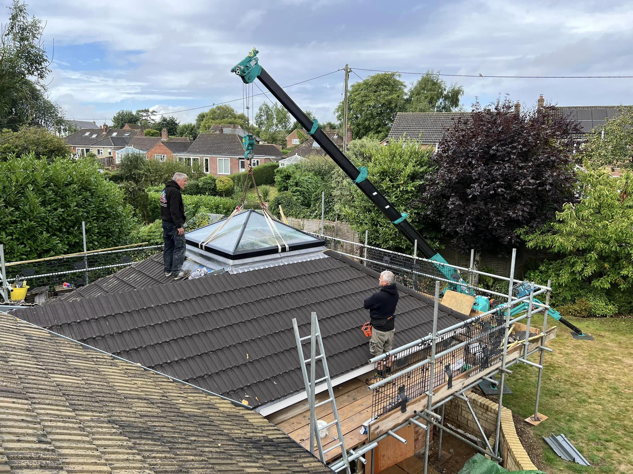 Two construction workers are installing a glass skylight on a pitched roof using a crane. The roof is surrounded by scaffolding, and the area is a residential neighborhood with trees, bushes, and houses in the background under a cloudy sky.