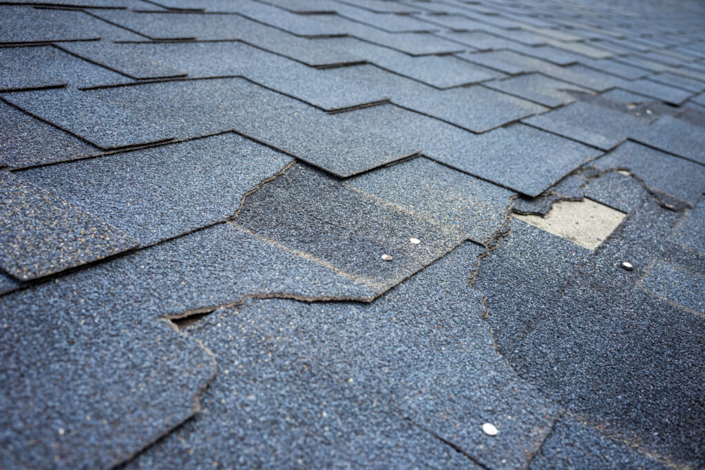 Close-up of a roof with worn, cracked, and damaged asphalt shingles. Several shingles are visibly curling and displaced, exposing the underlying material. Small nails are visible, securing some of the shingles in place. The roof appears to be in need of repair.