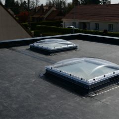 A flat rooftop with two dome-shaped skylights. The skylights are made of clear material framed with metal and are surrounded by a dark roofing material. In the background, there are several houses with slanted roofs, grassy yards, and some trees.