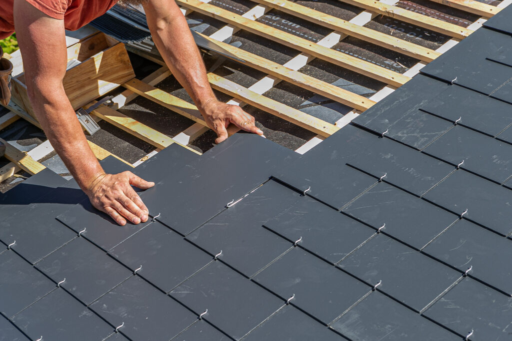 A person installing gray tiles on a roof. Their hands are placing a shingle onto the wooden framework, with some tools and materials visible in the background. The installation shows neat rows of interlocked tiles. The scene is lit by sunlight.