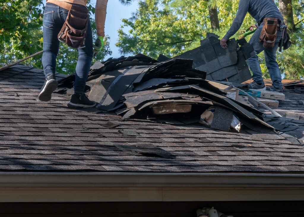 Two workers on a roof remove old shingles and replace them with new ones. One worker is seen from the waist down holding a tool, and the other is laying down shingles. They are surrounded by trees under a clear blue sky. Tools and materials are scattered on the roof.