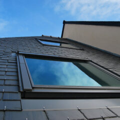 A low-angle view of a modern building with a tiled roof and large reflective windows against a blue sky with scattered clouds. The window panes reflect the sky, blending into the surrounding roofing material. The building's upper and side walls are visible.