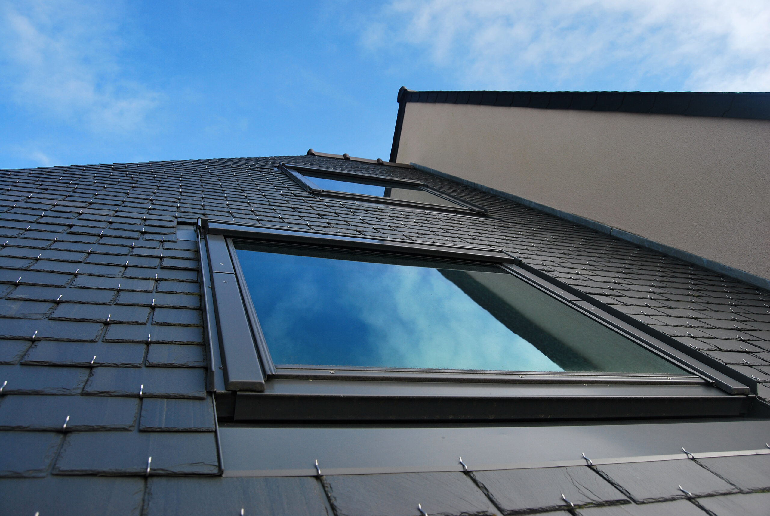 A low-angle view of a modern building with a tiled roof and large reflective windows against a blue sky with scattered clouds. The window panes reflect the sky, blending into the surrounding roofing material. The building's upper and side walls are visible.