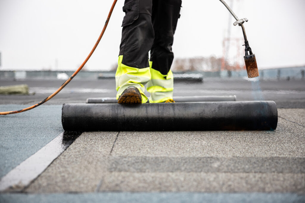 A worker in bright yellow safety gear is applying a waterproof membrane to a flat rooftop. They are using a gas torch to melt and seal the material while standing on a roll of the membrane. The background shows a blurred industrial setting.