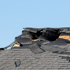 Image showing a close-up of a damaged roof with several shingles missing or curled up, exposing the underlying wooden structure. The damage appears to be significant, indicating the need for repair. The sky is clear and blue in the background.