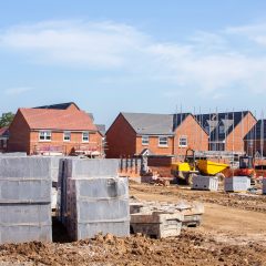 A construction site with several partially built red-brick houses. In the foreground, there are stacks of building materials, including pallets of bricks and concrete blocks. Scaffolding and construction vehicles are visible among the houses. The sky is clear.