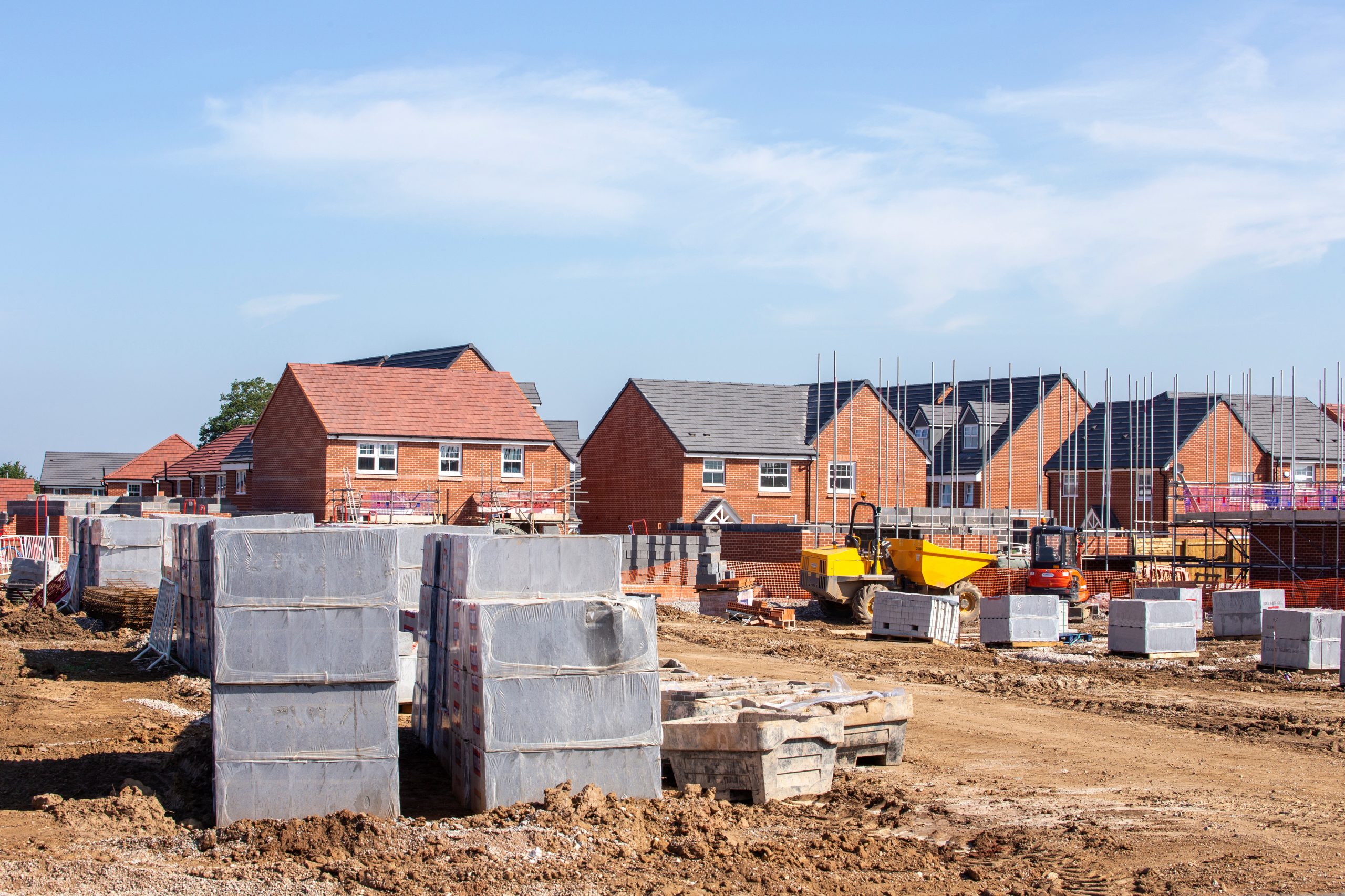 A construction site with several partially built red-brick houses. In the foreground, there are stacks of building materials, including pallets of bricks and concrete blocks. Scaffolding and construction vehicles are visible among the houses. The sky is clear.