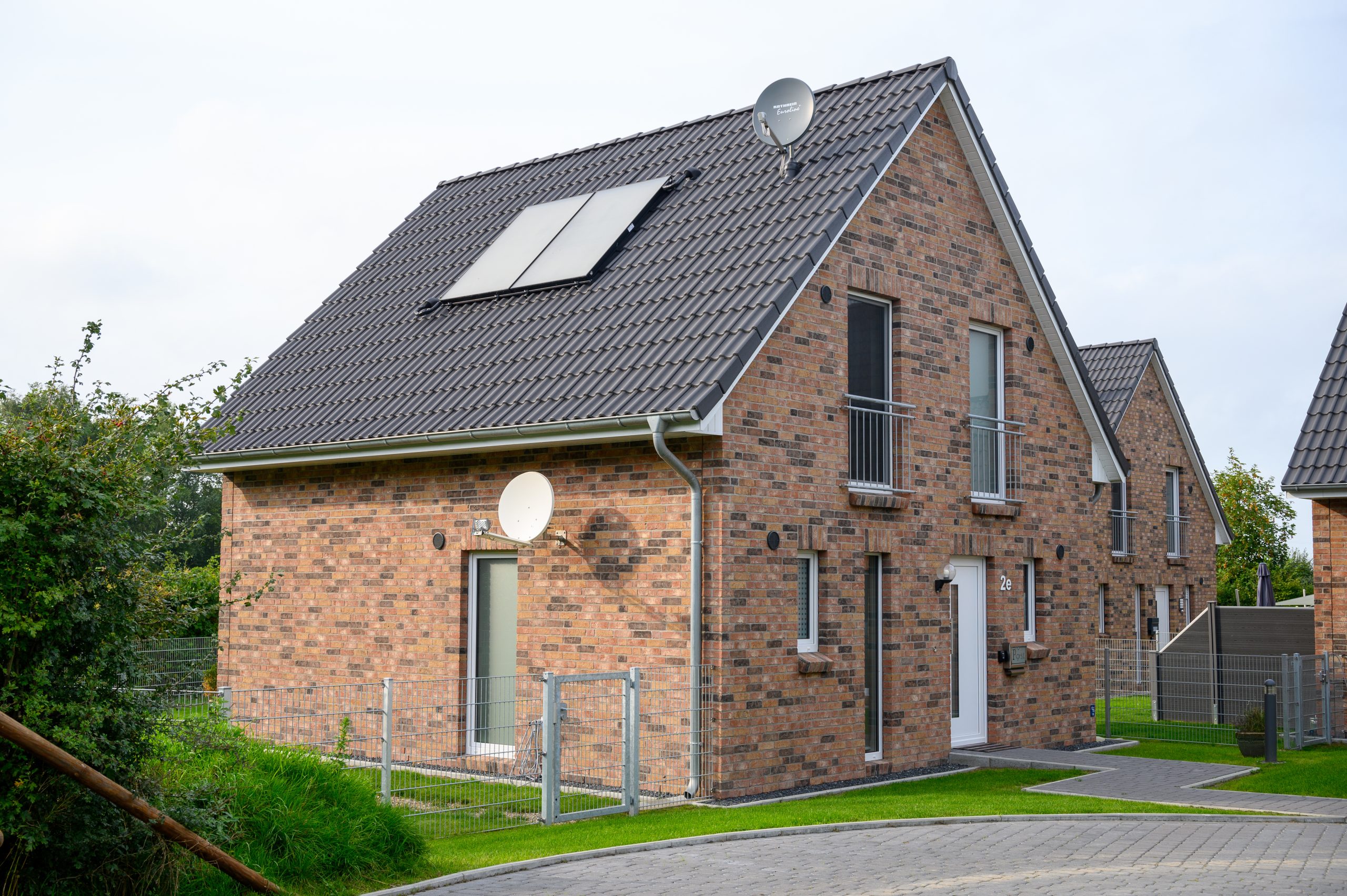 A modern brick house with a gabled roof featuring solar panels and satellite dishes. The house has large windows, a small balcony on the upper floor, and a gated entrance. The surrounding area has paved walkways and green lawns. Another similar house is in the background.