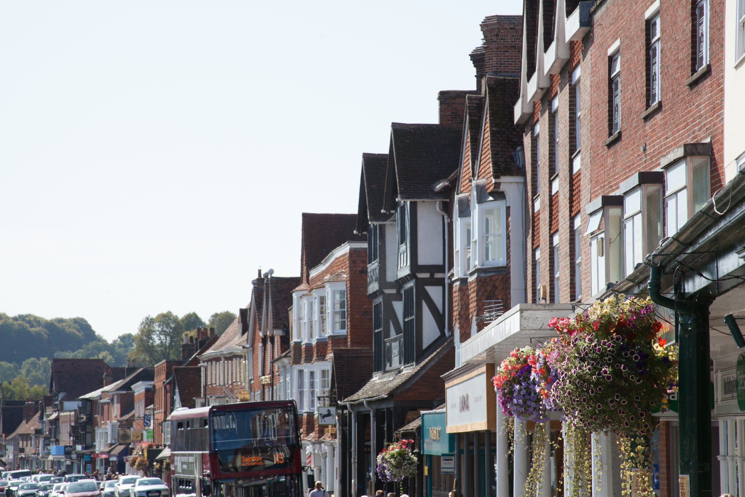 A busy street lined with various brick and timber-framed buildings, adorned with hanging flower baskets. A double-decker bus is visible on the left side of the street, and several people and cars are present, adding to the bustling atmosphere.