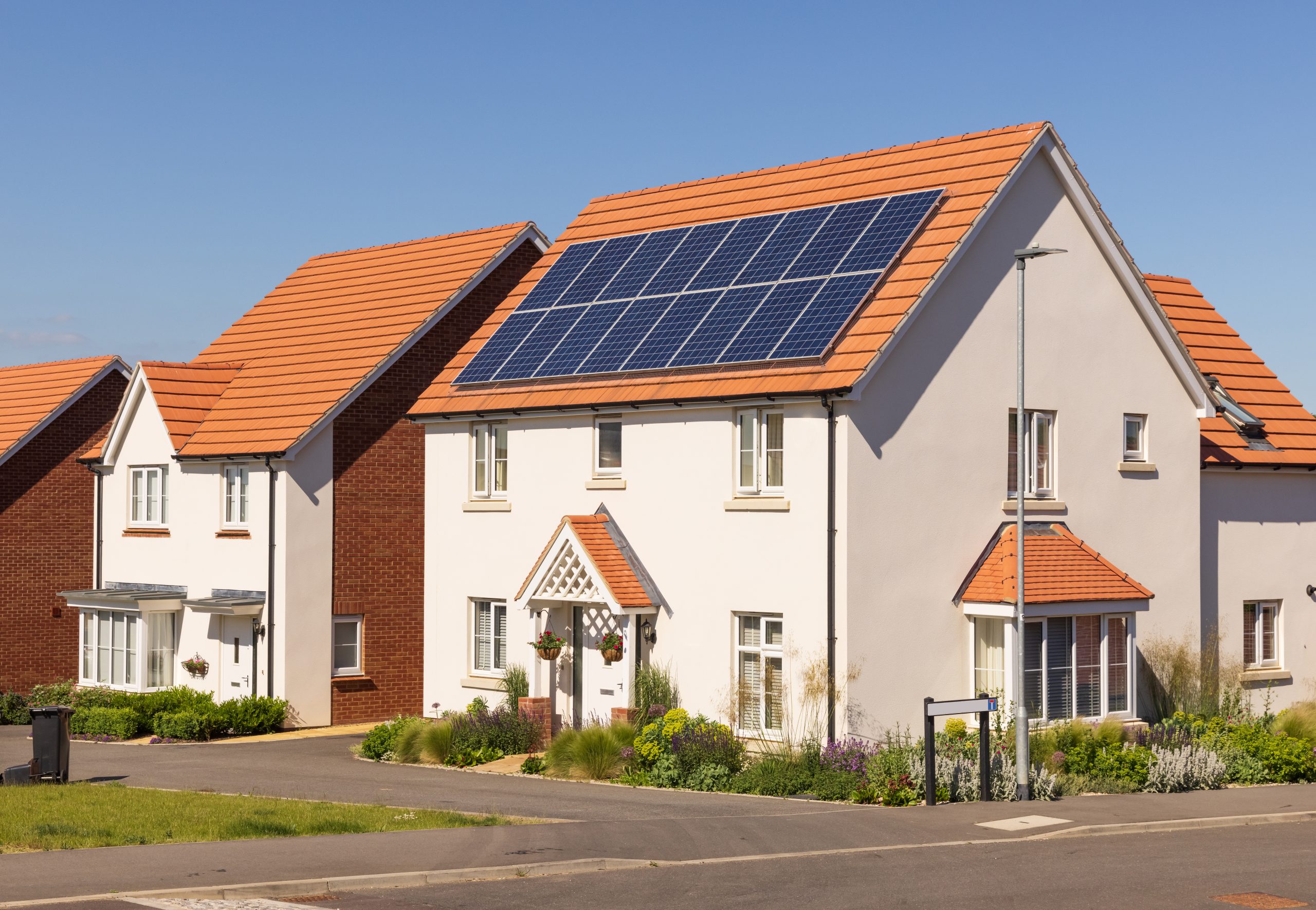 A row of modern, white houses with orange-tiled roofs under a clear blue sky. The house in the foreground has solar panels on its roof and a well-maintained front garden. The street is empty and clean.