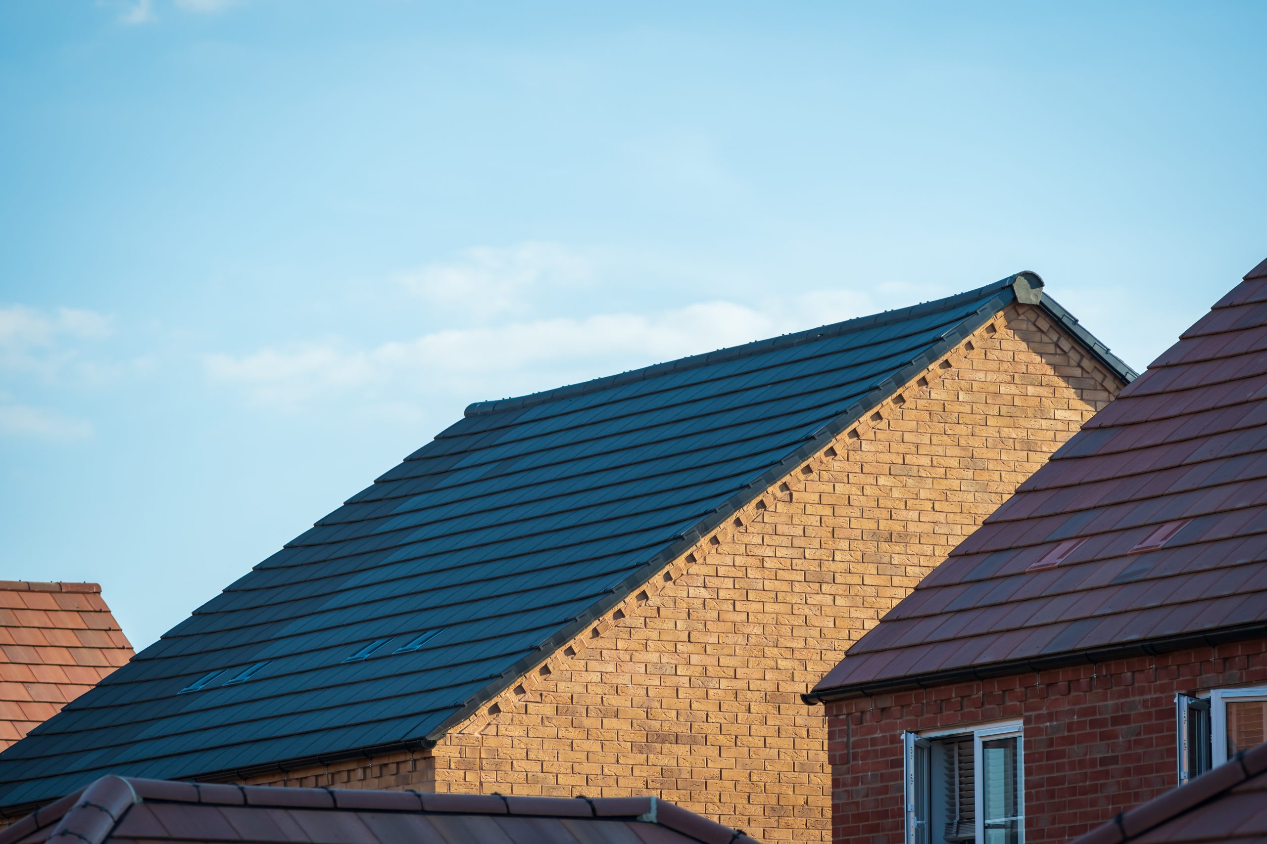 A close-up of several rooftops of brick houses under a clear blue sky. The central house features a light-brown brick facade with a dark blue slate roof. Other visible rooftops have reddish-brown shingles.