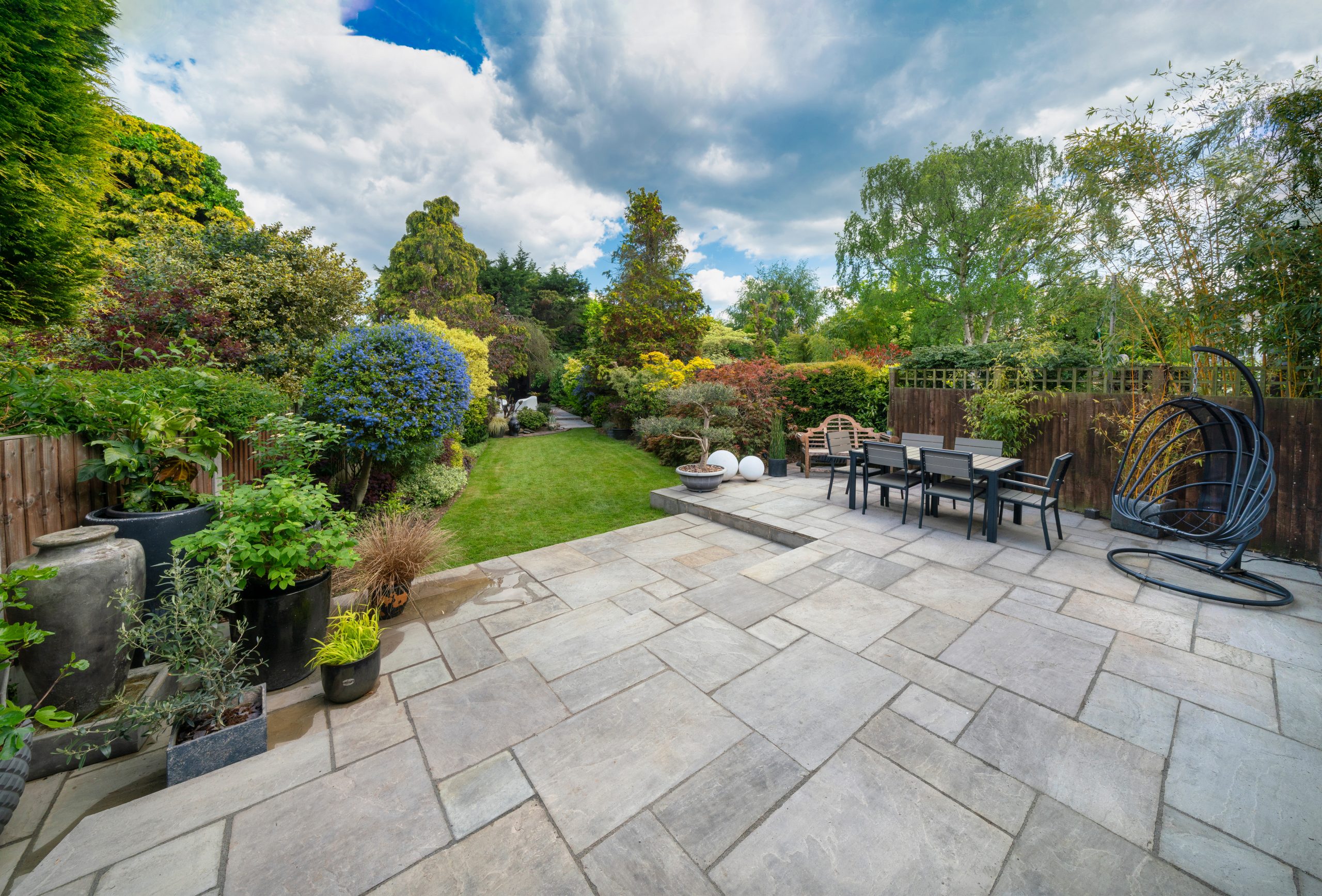 A spacious backyard with a well-landscaped garden features a large stone patio area. The patio includes a dining table with chairs and hanging chair swings. Vibrant flowers, various plants, and trees add bursts of color, extending into a lush green lawn.