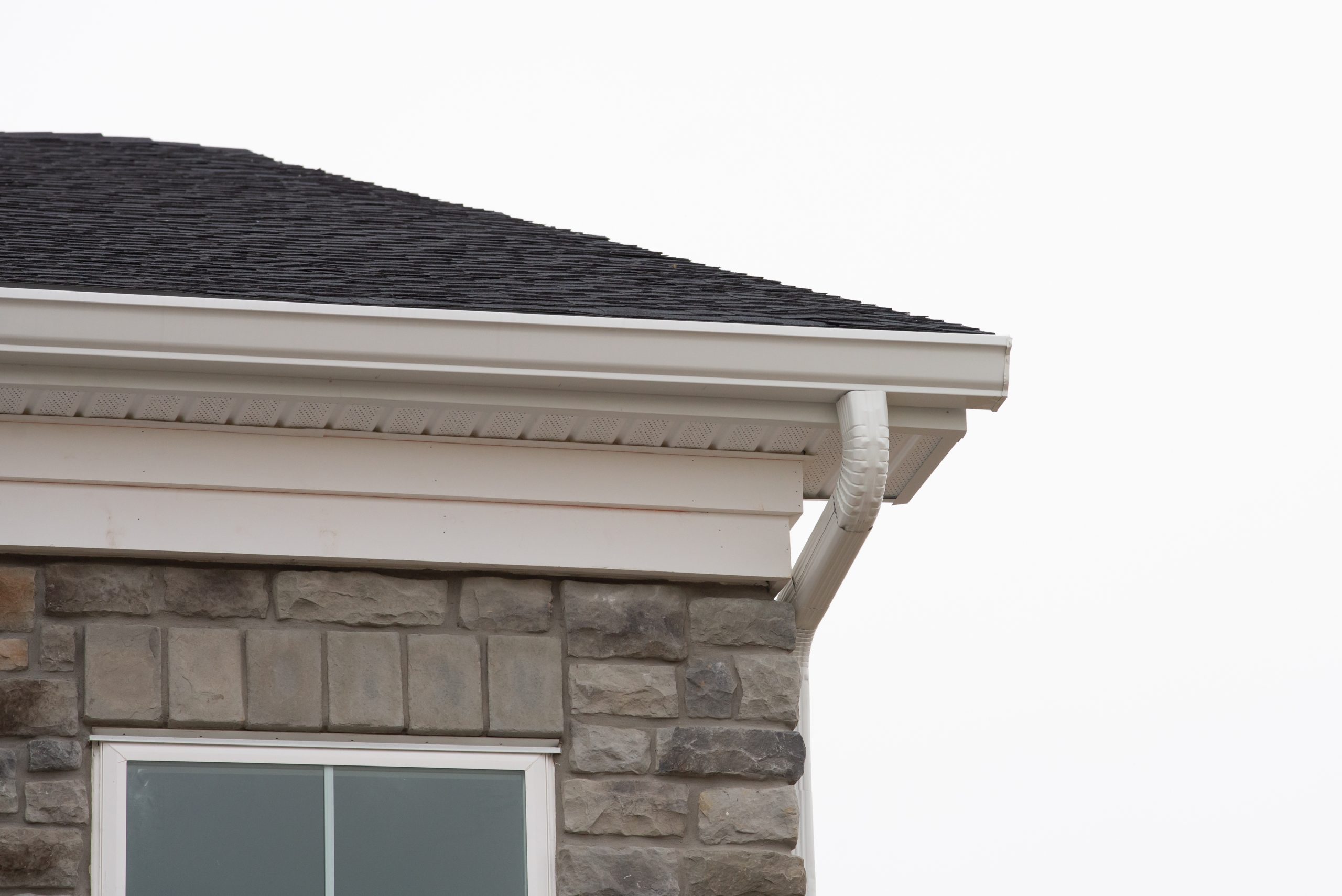 A corner of a house showcasing a stone exterior wall, a large window, and a black shingled roof. The roof features white gutters and downspouts. The sky in the background is overcast.