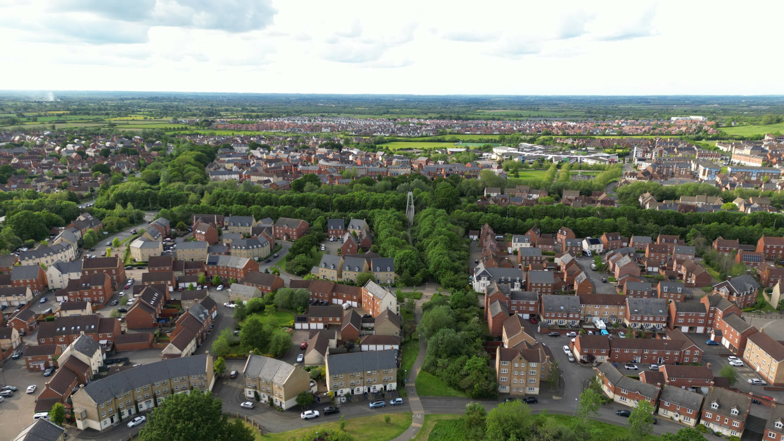 Aerial view of a suburban area with numerous houses lining curved streets, interspersed by green spaces. Trees form a significant cluster in the center, and fields and a distant horizon are visible in the background under a partly cloudy sky.