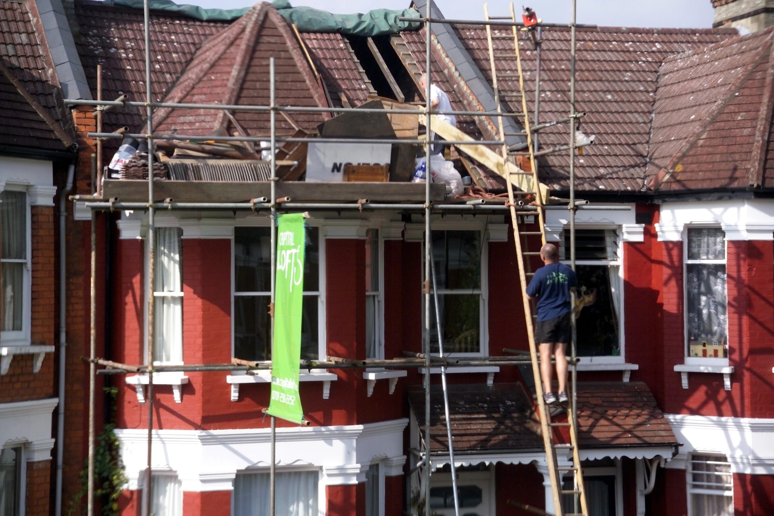 A worker stands on a ladder while renovating the roof of a red brick house with a white trim. Scaffolding surrounds the building, and another worker is visible on the roof. A bright green site sign is displayed in front of the house.