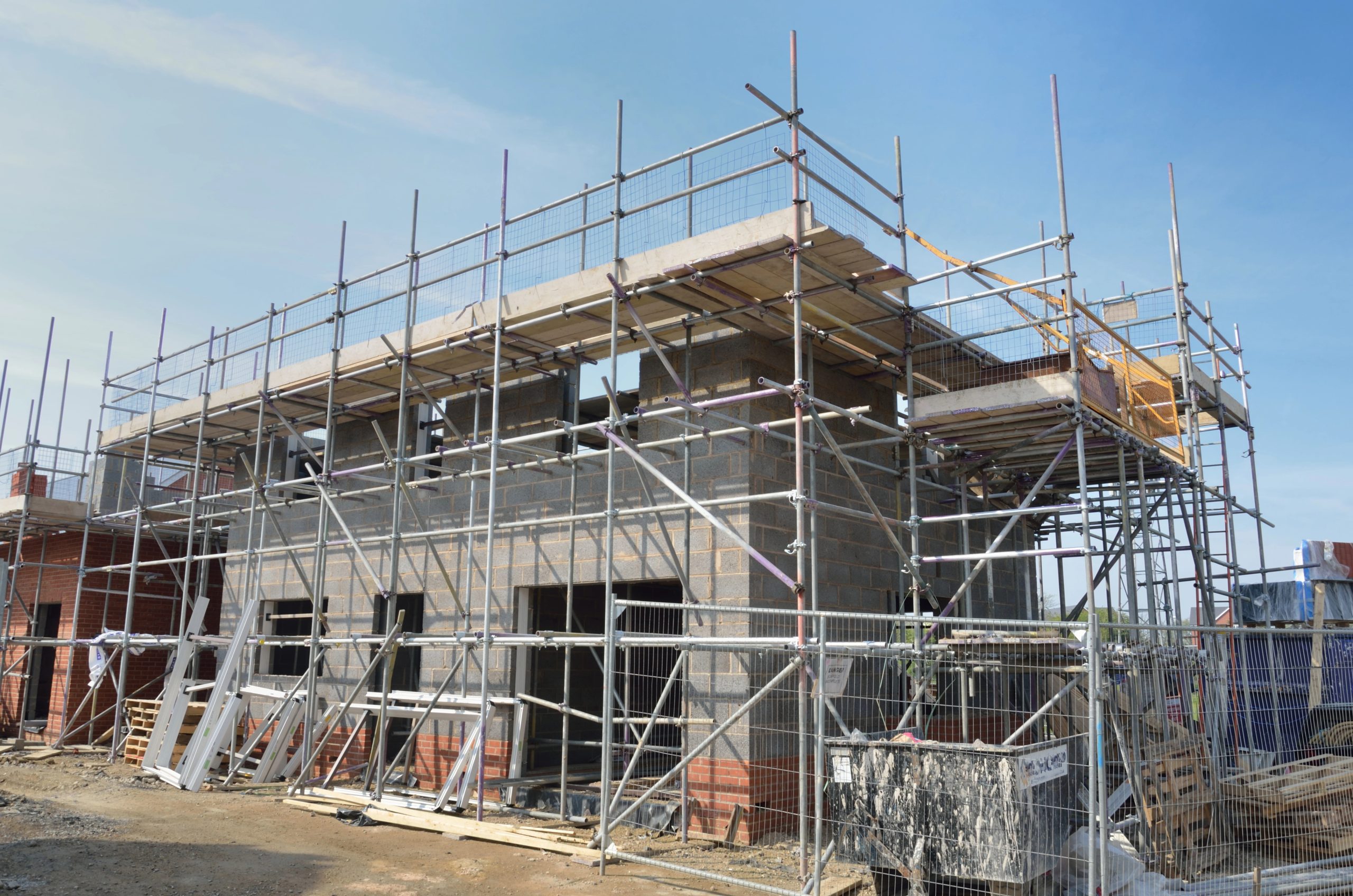 A partially constructed two-story brick building is surrounded by scaffolding and construction materials. The construction site is organized with metal scaffolding providing access to various levels of the building. The sky is clear and blue.