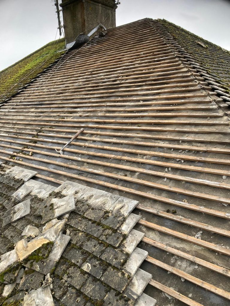 A sloped roof under renovation, showing exposed wooden battens and some missing tiles. A chimney is visible at the top, and a few remaining tiles lie askew. Moss is growing in patches on the remaining tiles.