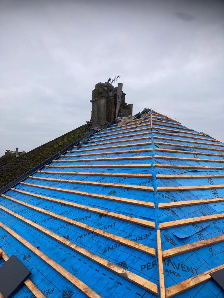 A roof under construction covered with blue tarp labeled "PERMAVENT APEX." Wooden battens are evenly spaced across the tarp. A chimney and some roof tiles are visible against a cloudy sky.