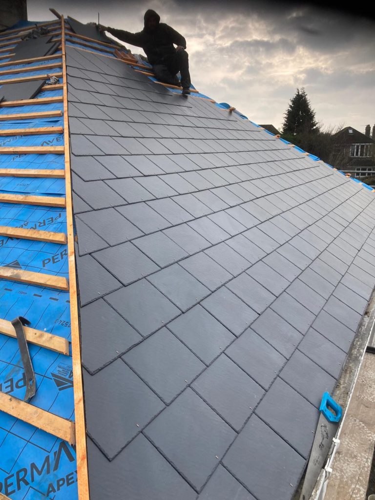 A roofer sits on a steep roof covered with new gray tiles. Wooden battens and blue roofing underlayment are visible on the unfinished section. A cloudy sky and a tree are in the background.