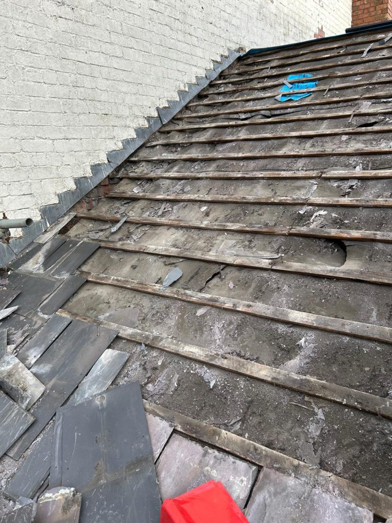 A sloped roof under repair, with exposed wooden beams and some missing or misplaced slate tiles. There is debris scattered around, and some blue and red objects are placed on the roof. A white brick wall is adjacent.