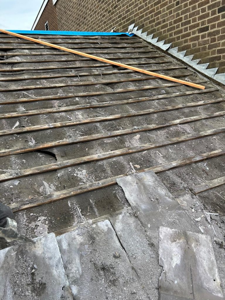 A close-up of a sloped roof under renovation. The old roofing material has been removed to expose wooden battens. A long wooden pole rests on the roof. Part of a brick wall is visible on the right.