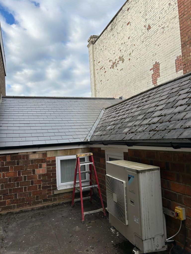 Brick building outdoor area with a red ladder leaning against the wall. Two roof sections meet, covered in slate tiles. An air conditioning unit is mounted nearby. The sky is partly cloudy, and some brickwork is worn.