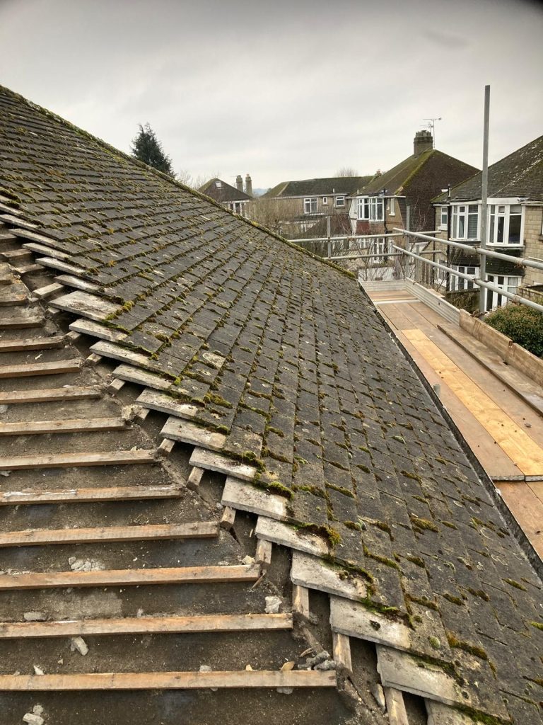 A sloped residential roof with old, moss-covered tiles, some of which are missing. Wooden battens are visible where tiles have been removed. Scaffolding surrounds the roof, set against an overcast sky and neighboring houses.