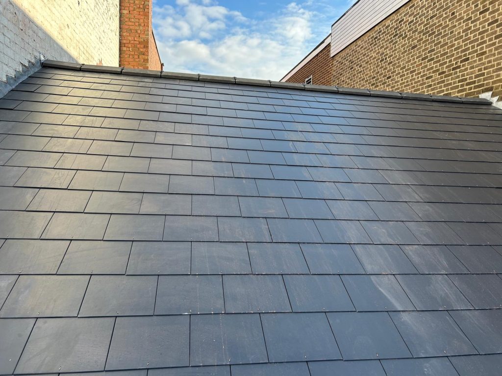 A newly constructed roof with dark slate shingles against a partly cloudy blue sky. The roof is bordered by brick walls on either side, showcasing a neat and modern design.