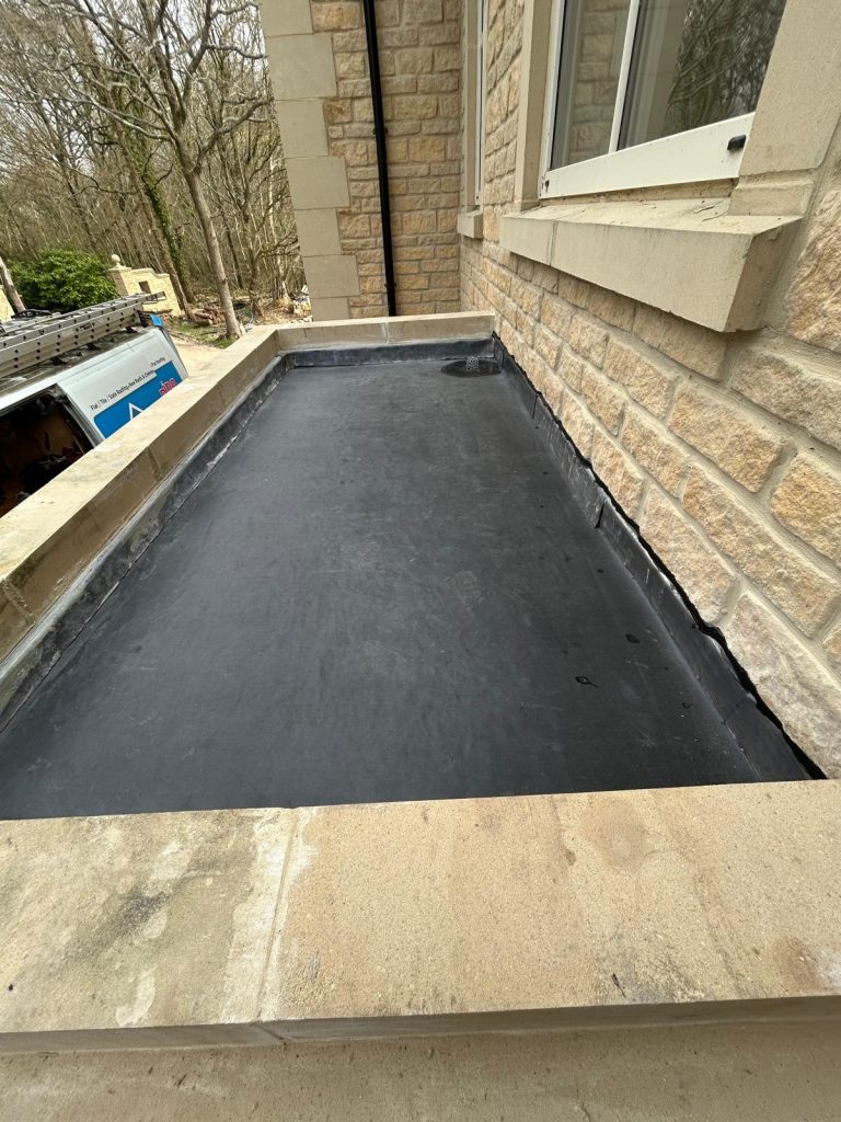 Flat stone ledge outside a brick building, sealed with black weatherproofing material. A window, part of the building, is visible on the right. Trees and construction materials are in the background.
