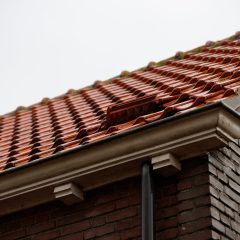 Close-up view of a sloped red-tiled roof with one displaced tile, showing part of a brick wall and rain gutter under an overcast sky.