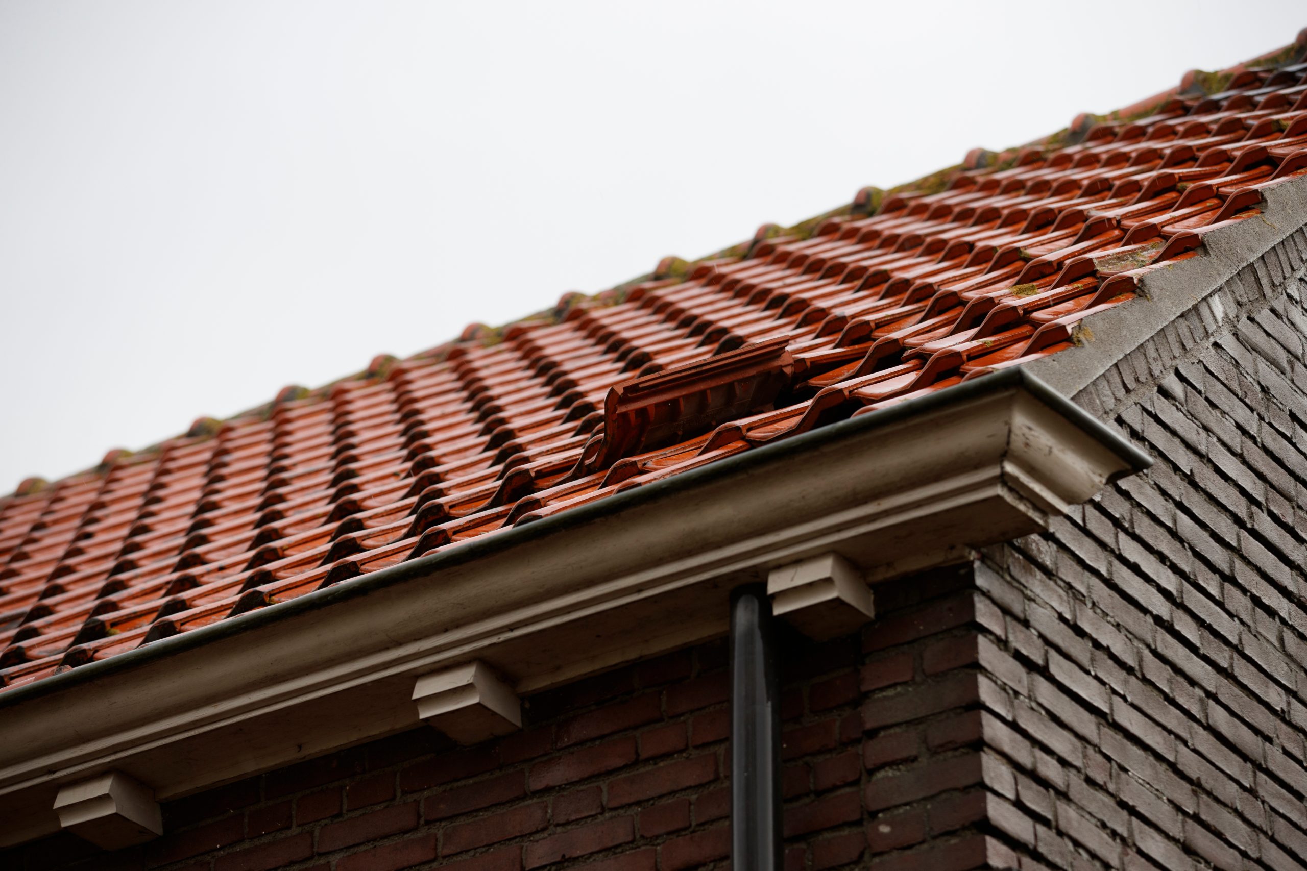 Close-up view of a sloped red-tiled roof with one displaced tile, showing part of a brick wall and rain gutter under an overcast sky.