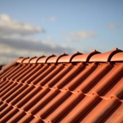 Close-up of a sloped roof with orange terracotta tiles under a blue sky with scattered clouds, photographed in warm sunlight.
