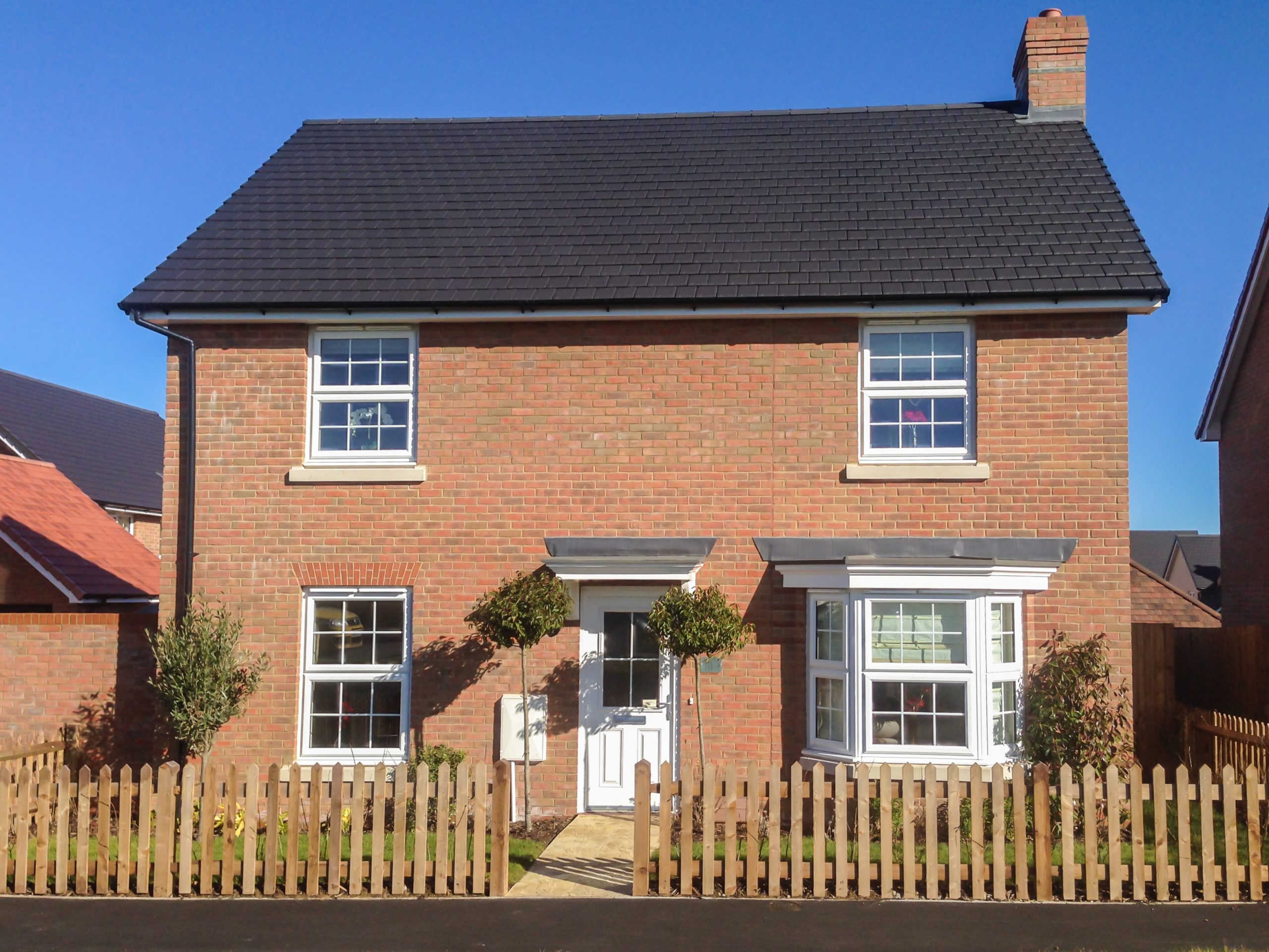 A modern two-story brick house with white trim and a dark roof, featuring a white front door, bay window, small trees, and a wooden picket fence, all under a clear blue sky.