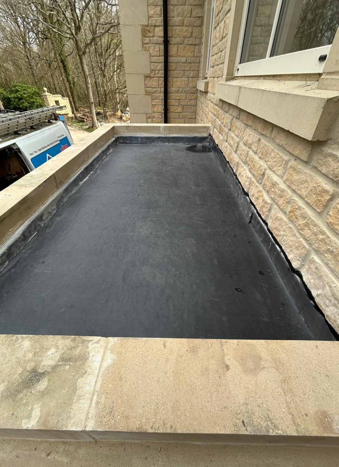 A flat, rectangular section of roof covered with black rubber material sits beneath a window on a stone building, with trees and a blue van visible in the background.