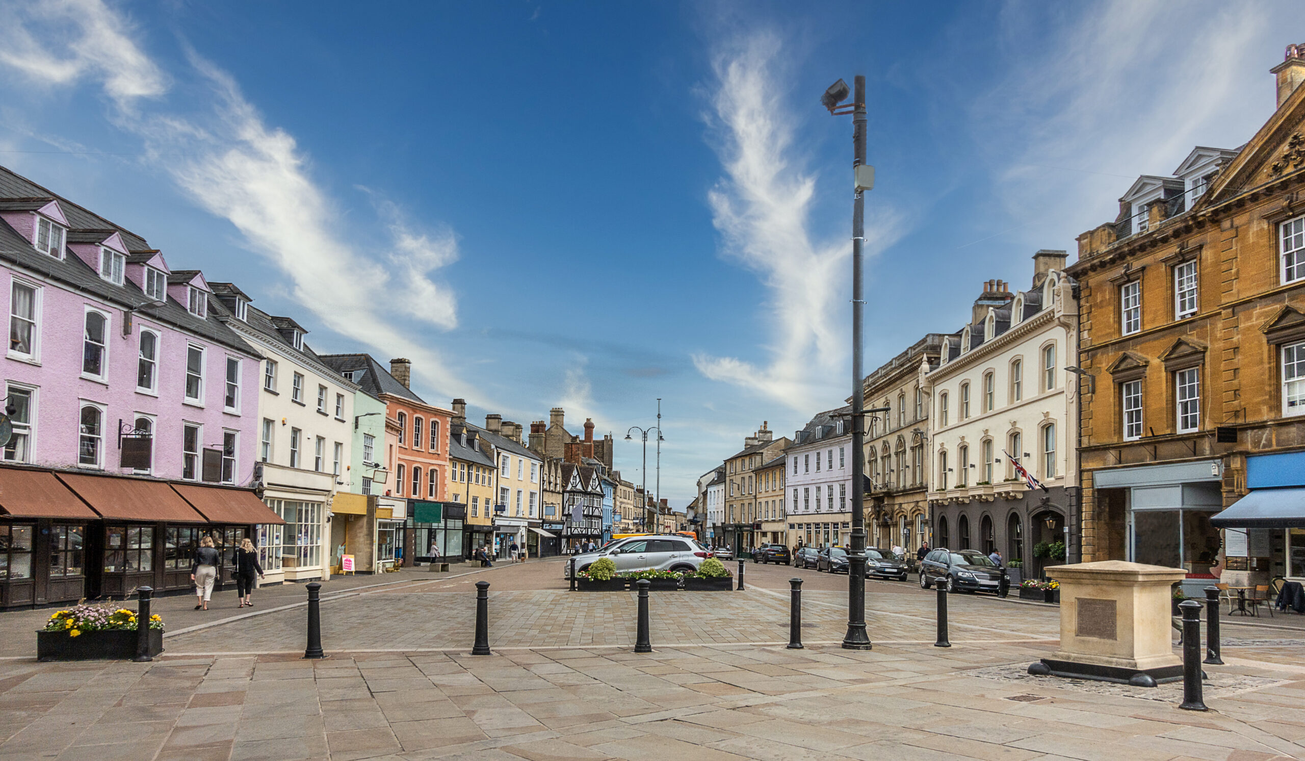 A wide cobbled street lined with colorful historic buildings, shops, and cafes under a blue sky with wispy clouds. Cars are parked along the sides, and a few people walk on the sidewalks.
