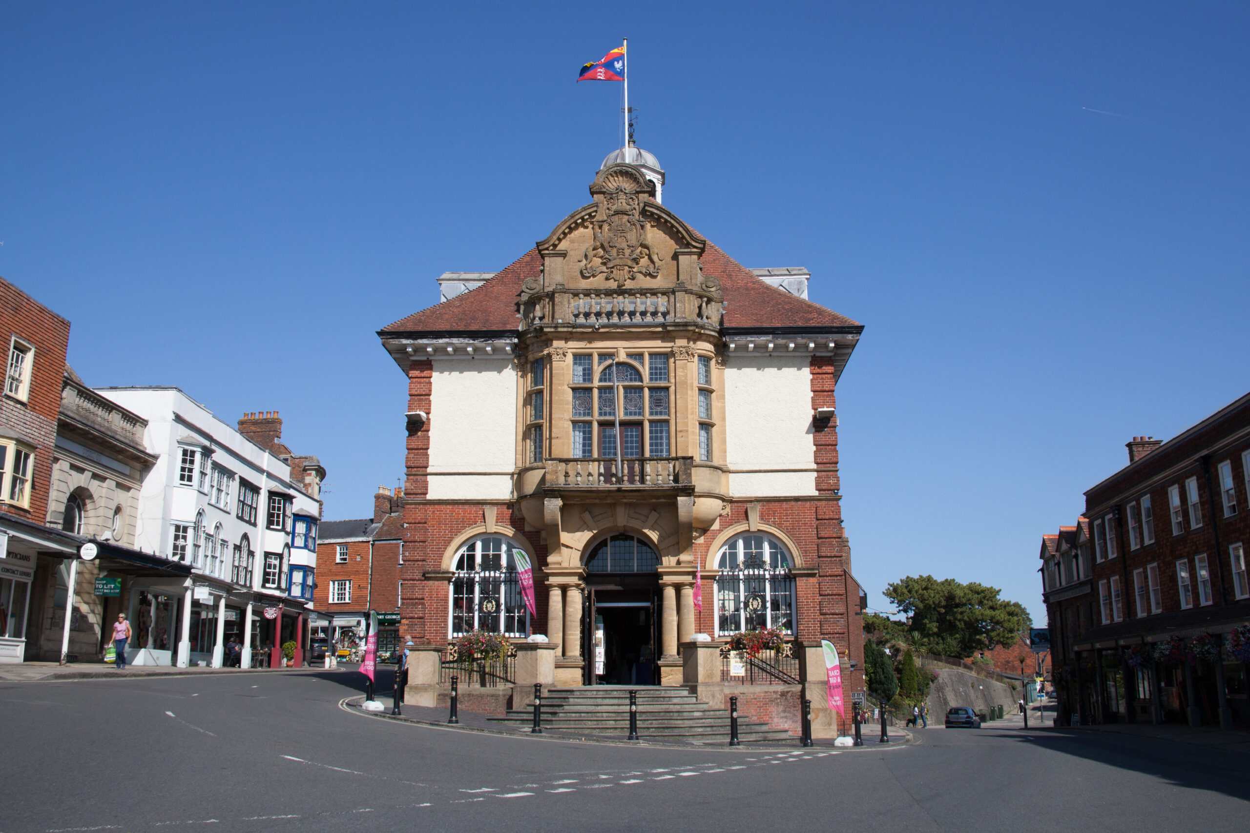 A historic brick building with an ornate balcony and flag on top stands at the corner of a quiet street with blue skies above. Other buildings line the street on both sides.