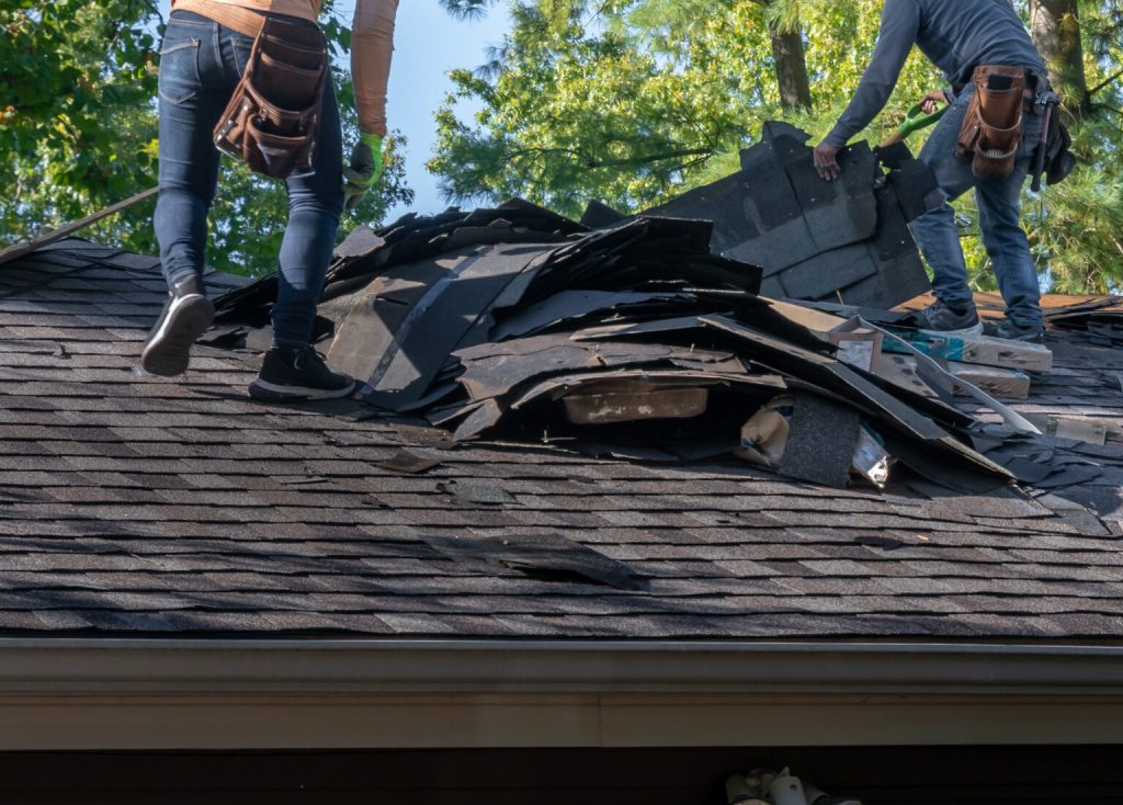 Two workers, wearing tool belts and jeans, remove old shingles from a house roof, creating a large pile of roofing debris. Trees with green leaves are visible in the background under a clear sky.