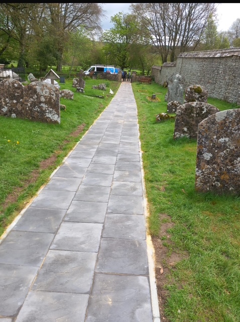 A paved walkway runs through a graveyard with old, moss-covered tombstones on both sides. Trees and grass surround the area, and a white and blue van is parked in the background near a stone wall.