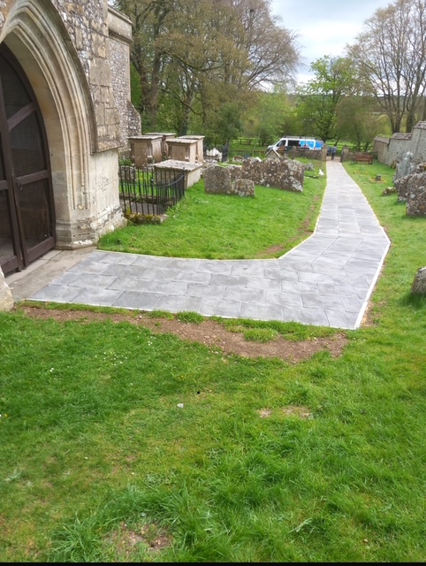 A paved path curves through a grassy churchyard beside an old stone building with arched doors, passing gravestones and trees, with a white vehicle in the background.