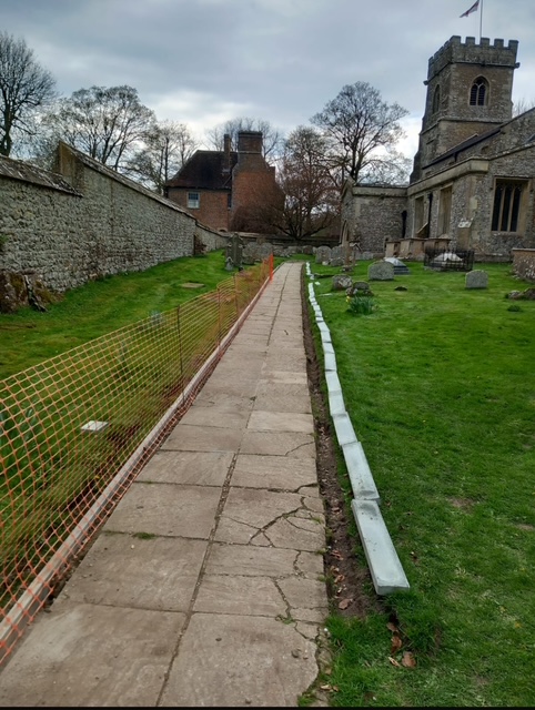 A stone path runs alongside a church with a tower, bordered by grass and graves on one side and an orange safety fence and stone wall on the other, under a cloudy sky.
